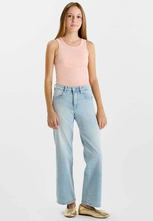 Tienermeisje staand, gekleed in een roze mouwloze top, lichtblauwe spijkerbroek en gouden ballerina's, tegen een witte achtergrond.