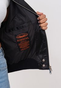 Zwarte bomberjack interieur met een oranje label waarop "BOMBERS ORIGINAL" staat, zilveren hardware en een gladde, satijnachtige textuur.