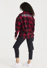 Camicia di flanella a quadri rossi e neri con testo bianco "SIKSILK" sulla schiena, abbinata a pantaloni cargo neri e sneakers bianche. Tessuto testurizzato.