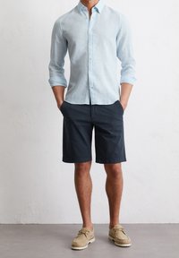 Uomo che indossa una camicia azzurro chiaro a maniche lunghe con maniche arrotolate, pantaloni pantaloncini blu scuro e scarpe casual beige, in piedi con le mani in tasca.