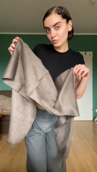 Lysbrun, tekstureret tørklæde holdt med begge hænder, draperet i luften, med frynset kanter. Bæres med en tætsiddende sort top og grå bukser.