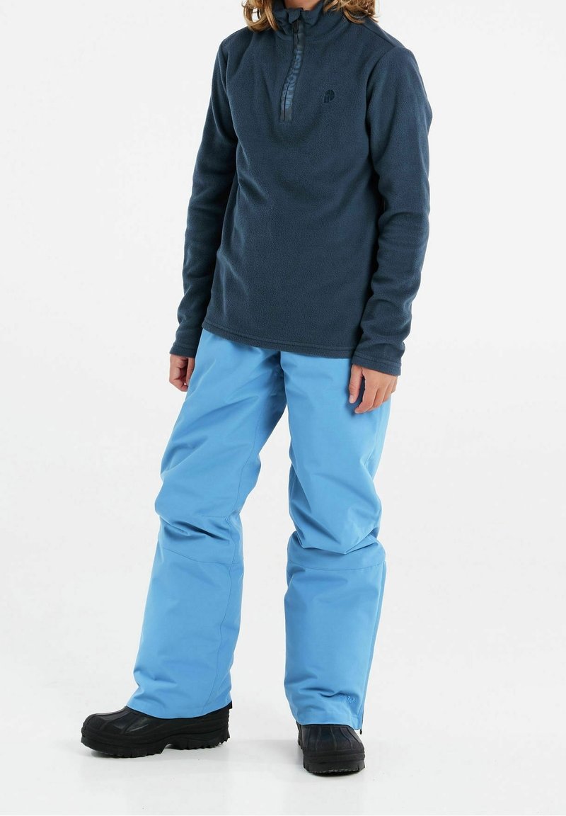 Enfant portant un pull polaire bleu foncé, un pantalon de neige bleu clair et des bottes d'hiver noires, debout sur un fond blanc.