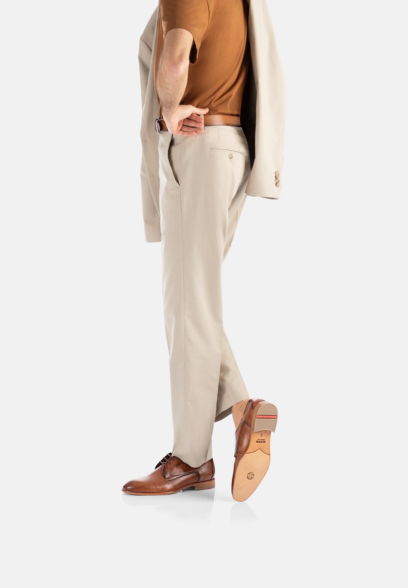 Bruine leren schoenen, beige broek en een bruine shirt; persoon houdt een lichtgekleurde jas vast met een slim fit en maatwerk ontwerp.