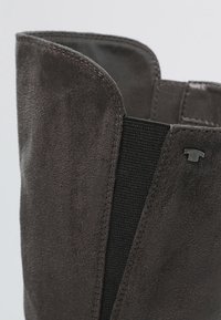 Gros plan sur la tige d'une botte en daim marron foncé avec un panneau latéral élastique et une petite étiquette métallique avec logo près du bord supérieur.