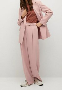 Světlounce růžový oblek se sestává z elegantního saka na míru a kalhot s širokými nohavicemi, doplněný tmavým topem a bílými teniskami. Hladká textura a pohodlný střih.