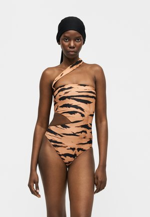 Slank kvinde iført en tigerstribet badedragt med én skulder og en udskæring i siden samt et sort tørklæde på hovedet mod en ensfarvet baggrund.