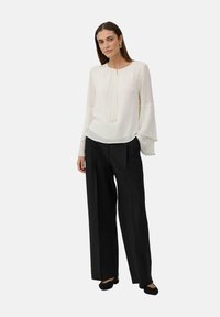 Femme portant un chemisier blanc à manches longues avec des poignets superposés, un pantalon noir à jambes larges et des chaussures noires, se tenant avec une main dans la poche.