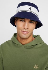 Chapeau bob rayé bleu marine et blanc avec une texture douce, arborant un accent de logo. Porté par une personne vêtue d'un hoodie vert olive.