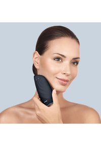 Svart ansiktsrengöringsapparat med en böjd, texturerad yta, hålls mot huden. Slät yta, kompakt form och varumärkeslogotyp synlig.