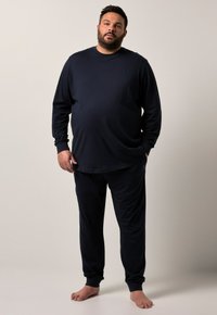 Man som står barfota iförd en svart långärmad tröja och svarta byxor mot en enkel beige bakgrund.