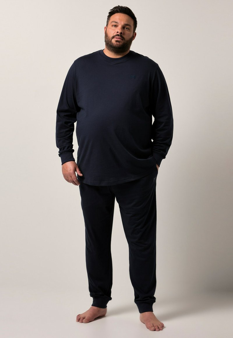 Man som står barfota iförd en svart långärmad tröja och svarta byxor mot en enkel beige bakgrund.