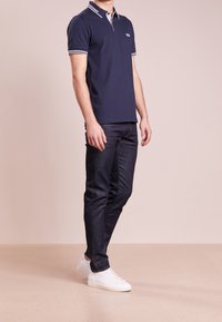 Polo shirt marine avec des bordures blanches et grises, à manches courtes, porté avec un jean foncé et des baskets blanches, arborant un logo sur la poitrine.