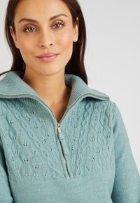 Maglione azzurro chiaro a maglia con colletto con zip, caratterizzato da un motivo testurizzato nella parte superiore e un tessuto liscio nella parte inferiore. Dettagli in zip dorati.