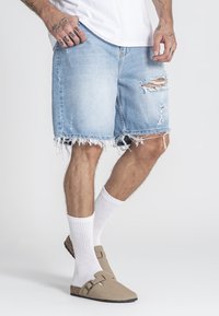 Svijetloplavi iznošeni trapke s franjastim rubovima, nošeni s bijelim čarapama do gležnja i smešim natikačama s remenom.