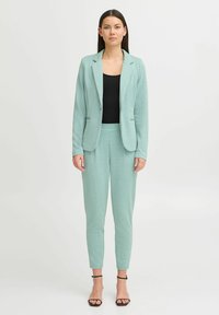 Lichtgroene blazer en aansluitende broek in een gestructureerde stof, gecombineerd met een zwart top. Voorzien van een open voorkant en een slanke snit.