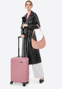 Roze harde koffer met geribbelde textuur, zwarte handgreep en wielen, gecombineerd met een zwarte leren jas en een roze schoudertas.