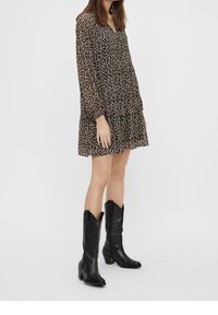 Robe noire à motif floral avec manches longues et col en V ; design superposé et fluide. Associée à de hautes bottes de cowboy en cuir noir, avec un bout pointu.