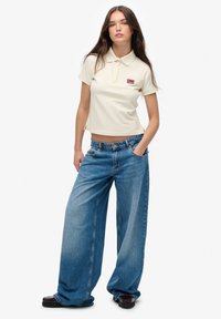 Lichtbeige polo shirt met korte mouwen en een klein logo. Losvallende blauwe denim jeans met een hoge taille en wijde benen. Zwarte schoenen.