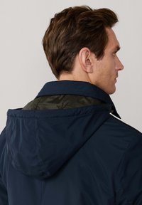 Marineblaue Kapuzenjacke mit einer glatten Außenseite, die ein kontrastierendes grünes Innenfutter und einen Kragen aufweist. Minimalistisches Design mit einem Reißverschluss.