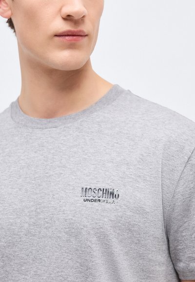 Νεαρός άνδρας που φοράει ανοιχτό γκρι μπλουζάκι με το λογότυπο "MOSCHINO UNDERWEAR" κεντημένο στο στήθος, στραμμένος ελαφρώς προς τα δεξιά.