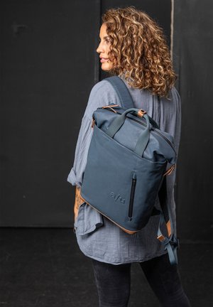 Navyblaue Rucksack mit einem Rollverschluss, Lederakzenten, Frontreißverschlussfach und verstellbaren Trägern, auf einem Modell präsentiert.