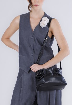 Femme portant un gilet gris sans manches boutonné et un pantalon, tenant un sac à main en cuir noir avec un détail floral sur la lanière et le gilet.
