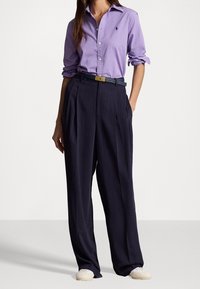 Camisa de manga larga de color lila claro con botones, combinada con pantalones de talle alto azul marino oscuro con pliegues. Los accesorios incluyen un cinturón oscuro.