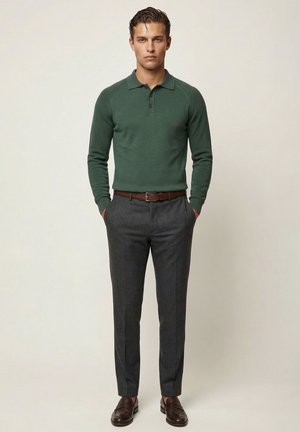 Mand står med hænderne i lommerne iført en grøn langærmet polo, grå bukser, brun bælte og brune læder loafers mod en neutral baggrund.