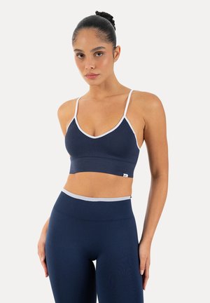 Młoda kobieta z upiętymi włosami, ubrana w granatowy sportowy biustonosz i legginsy z wysokim stanem z białą lamówką, stoi na jednolitym tle.