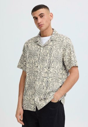 Mand med kort hår iført en kortærmet cremefarvet skjorte med sorte blomsterprint og sorte bukser, poserer med den ene hånd i lommen.