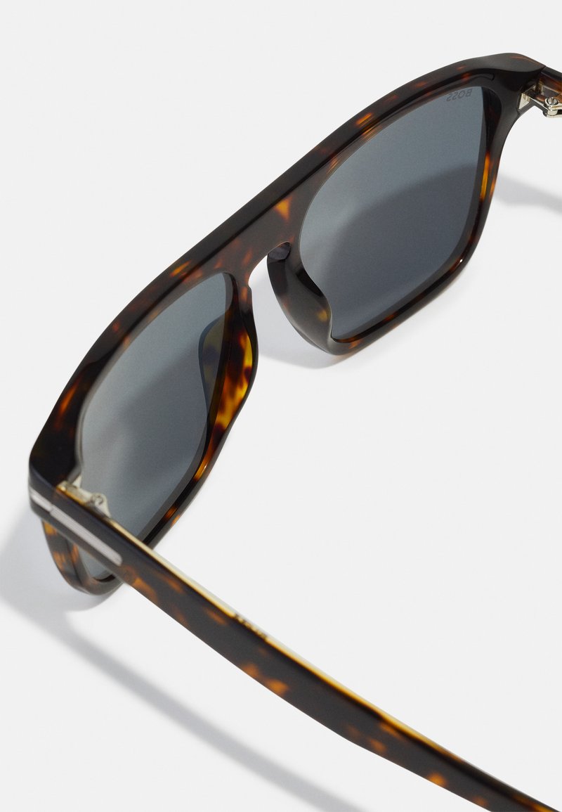 Occhiali da sole tartarugati con lenti grigie. Montatura spessa e angolare con una finitura lucida e un sottile accento dorato sulla stanghetta.