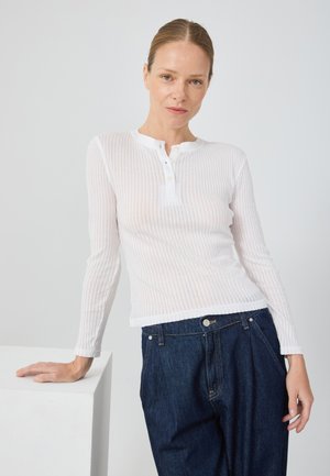 Weißes, geripptes Langarmoberteil mit Henley-Ausschnitt und Knopfdetails, kombiniert mit dunkelblauen, hochgeschnittenen Jeans. Glatte Textur.