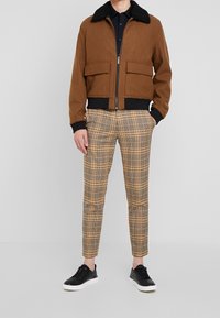 Bruin wollen bomberjack met zwarte kraag, gecombineerd met geel-bruin geruite broek en zwarte sneakers. Voorzien van rits en zakken aan de voorkant.