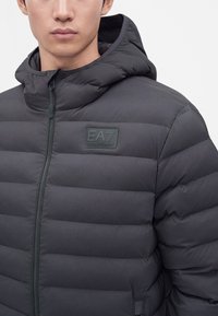 Άνδρας που φοράει μαύρο καπιτονέ μπουφάν EA7 Emporio Armani με κουκούλα και φερμουάρ στο μπροστινό μέρος, σε απλό φωτεινό φόντο.