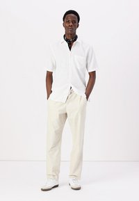 Camicia bianca a maniche corte con bottoni, pantaloni beige chiaro, sneakers bianche con suole color gomma e un bandana nera. Design semplici, vestibilità rilassata.