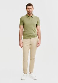 Oliwkowa koszulka polo z krótkim rękawem, w połączeniu z beżowymi chinosami o slim fit i białymi sneakersami, na jednolitym białym tle.