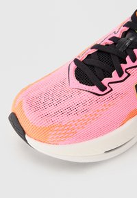 Baskets de sport en mesh rose et orange avec des lacets noirs, bout rond et semelle texturée. Comprend un logo sur le côté et un motif en grille.