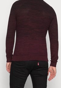 Mörkröd stickad tröja med ett texturerat mönster, ribbade muddar och nederkant. Svarta jeans med en röd logotag synlig på bakfickan.