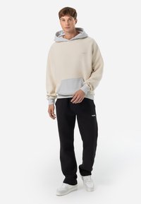 Beige fleece hoodie met een lichtgrijze capuchon en een kangoeroezak, gecombineerd met zwarte joggingbroek en witte sneakers. Eenvoudig, comfortabel ontwerp.
