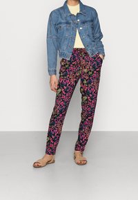 Jeansjacka med knappar, gul topp och svarta blommiga byxor med rosa, orange och gröna mönster. Beige sandaler fullbordar looken.