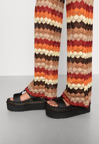 Zwarte platform sandaal met een dikke rubberen zool; gecombineerd met een multicolore gebreide broek met golfpatronen in oranje, bruin en roomkleur.