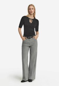 Svart top med kort ärm och nyckelhål-design samt knytdetalj, tillsammans med ljusgrå vida byxor och svarta skor.