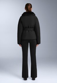 Žena s smeđom konjskom repom stoji okrenuta leđima, nosi crnu jaknu s pojasom i ovratnikom od krzna te crne hlače s patentnim zatvaračima na stražnjoj strani.