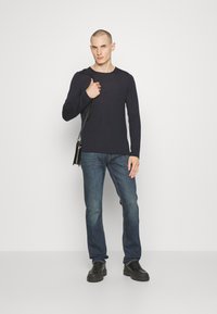 Camicia blu navy a maniche lunghe, slim fit, abbinata a jeans blu a gamba dritta. Il modello tiene una borsa a tracolla nera e indossa scarpe nere. Sfondo bianco.