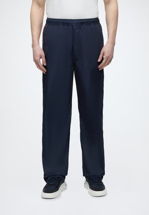 Man draagt marineblauwe, loszittende broek met een elastische tailleband en zwarte sneakers tegen een effen witte achtergrond.
