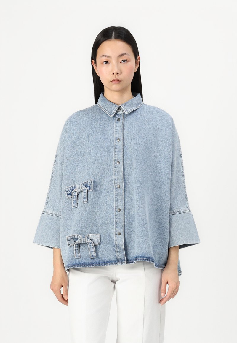 Ljust blå denimskjorta med krage, tryckknappar och tre dekorativa tygrosetter. Har vida ärmar och en lös passform.