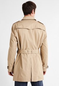 Mężczyzna w beżowym trenczu z paskiem i zapinanymi mankietami, zwrócony plecami na tle jednolitego białego tła.