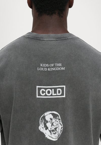 Persoon in een vervaagde grijze shirt met witte tekst "KIDS OF THE LOUD KINGDOM," een omkaderd "COLD," en een afbeelding van een schreeuwend gezicht op de achterkant.