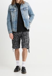 Person i ljusblå denimjacka, svart T-shirt, grå kamouflage cargo-shorts, vita strumpor och svarta tofs-loafers, stående mot vit bakgrund.
