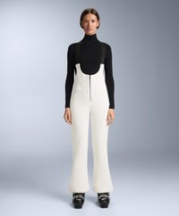 Białe dziane ogrodniczki z teksturowanym wzorem i regulowanymi czarnymi paskami, połączone z czarną bluzką z długim rękawem i czarnymi butami narciarskimi.